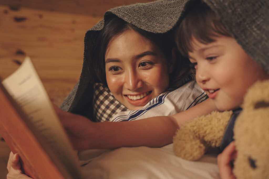 A mother and her preschool-age child snuggle under a blanket while reading a bedtime story, both smiling with joy. This cozy scene illustrates early reading routines for preschoolers and the role of storytime in nurturing literacy, imagination, and parent-child connection.