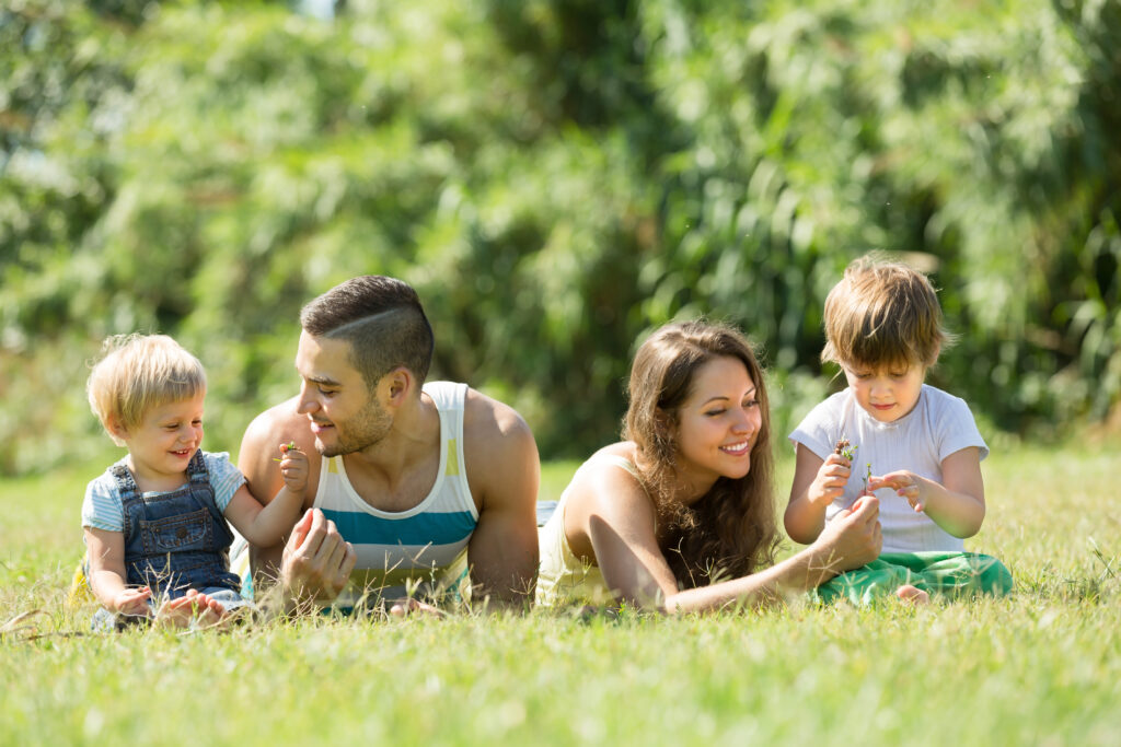 Parents and their two young children lie in the grass, smiling and playing with flowers under a sunny sky. This playful, peaceful moment captures the spirit of slow, joyful summer routines for kids