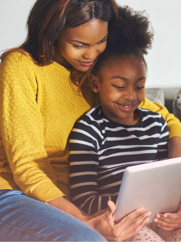 A mother and daughter sit close together on a couch, smiling as they look at a tablet, showing how parents can support online learning through shared screen time.