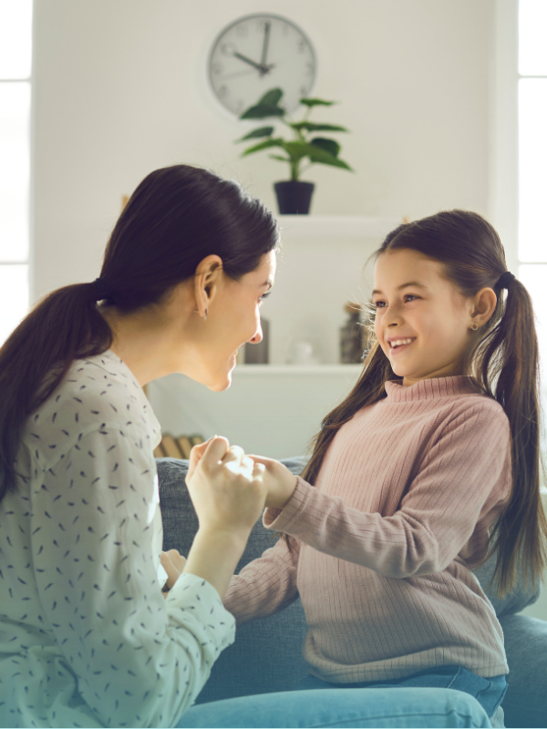 Mother and daughter smiling and holding hands while sitting on a couch, engaging in a warm and positive conversation at home.