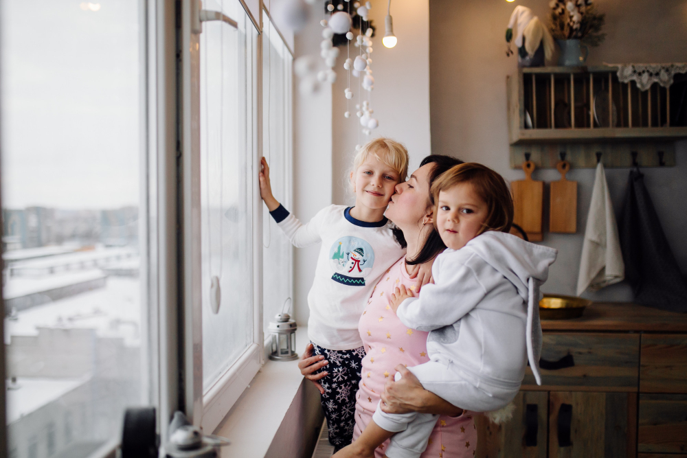 A mother lovingly kisses one of her two young children while standing by a bright kitchen window on a snowy day, symbolizing warmth, connection, and the power of self-care routines that strengthen families.